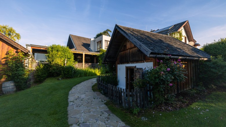 A small garden path leads to a traditional wooden house with flowers and bushes, surrounded by other buildings under a blue sky.