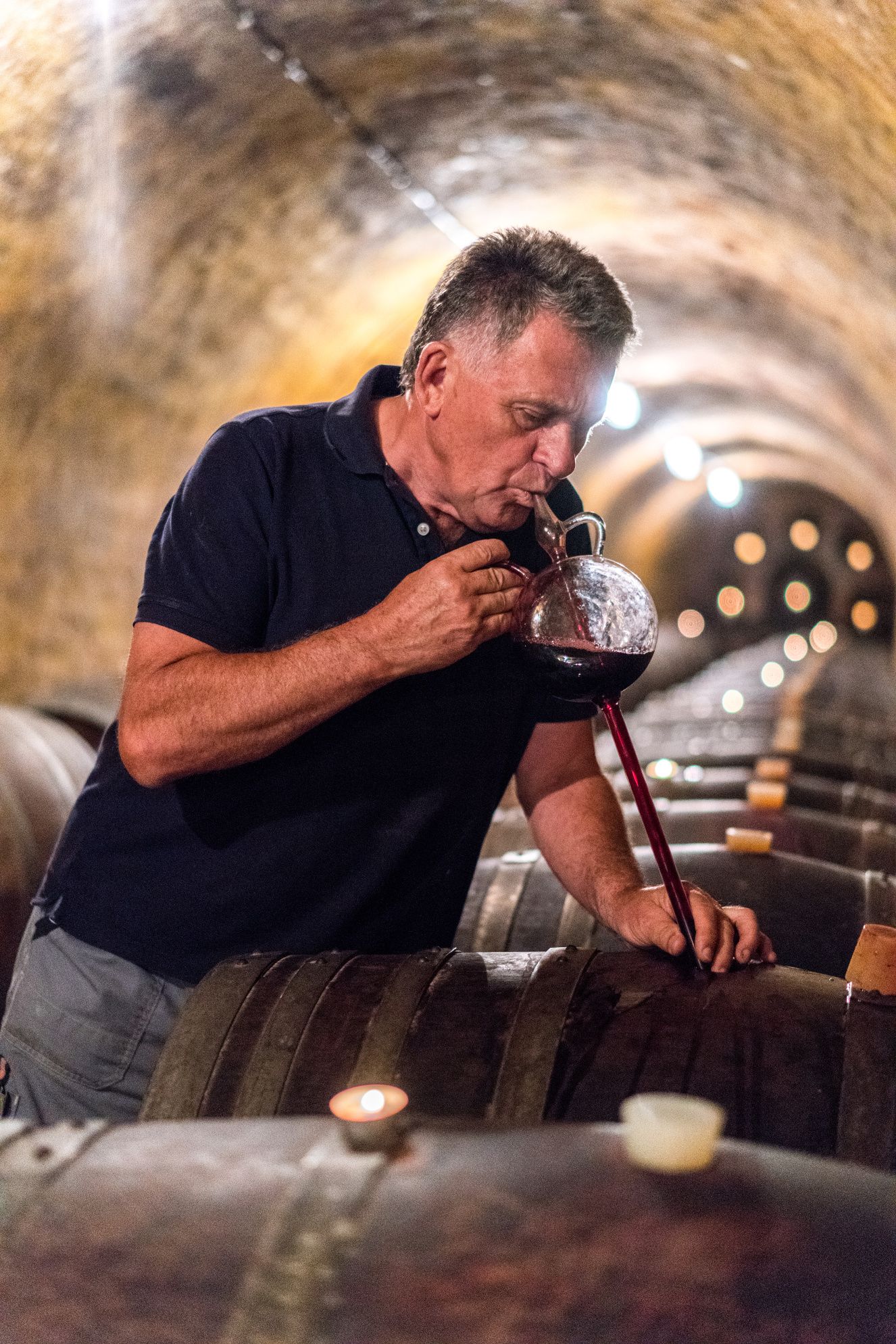 A man tastes wine from a barrel in a wine cellar.