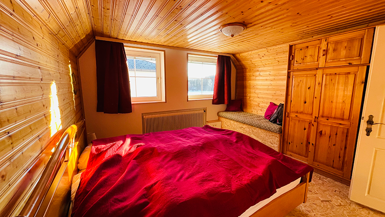 Wood-paneled bedroom with red comforter cover, two windows, a wardrobe and a bench.