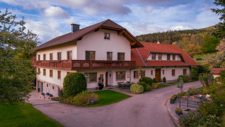 A traditional farmhouse with a red roof and balcony, surrounded by green countryside and trees.