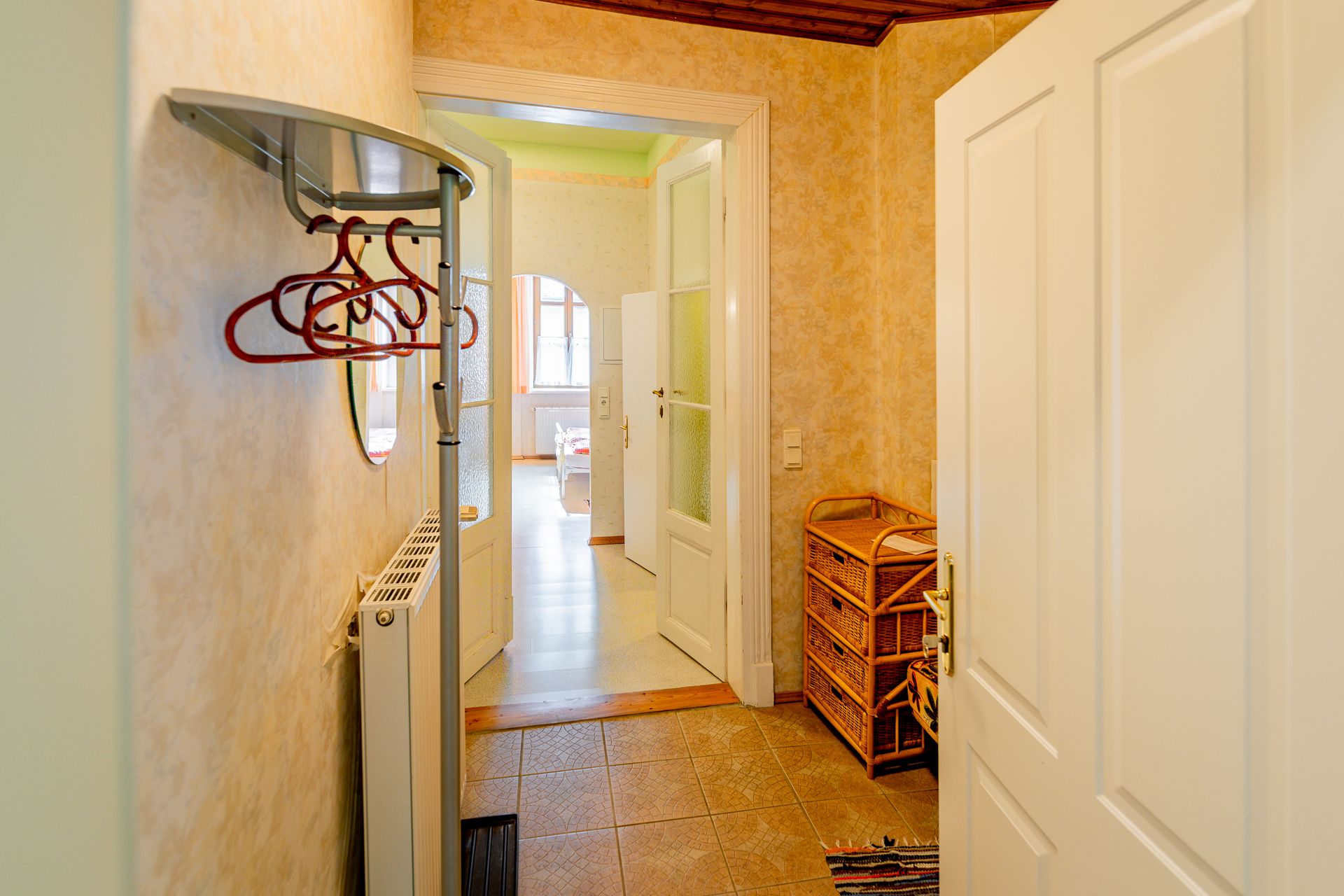 An anteroom with coat hooks, a radiator and wicker furniture. A door leads into a bright room with windows.