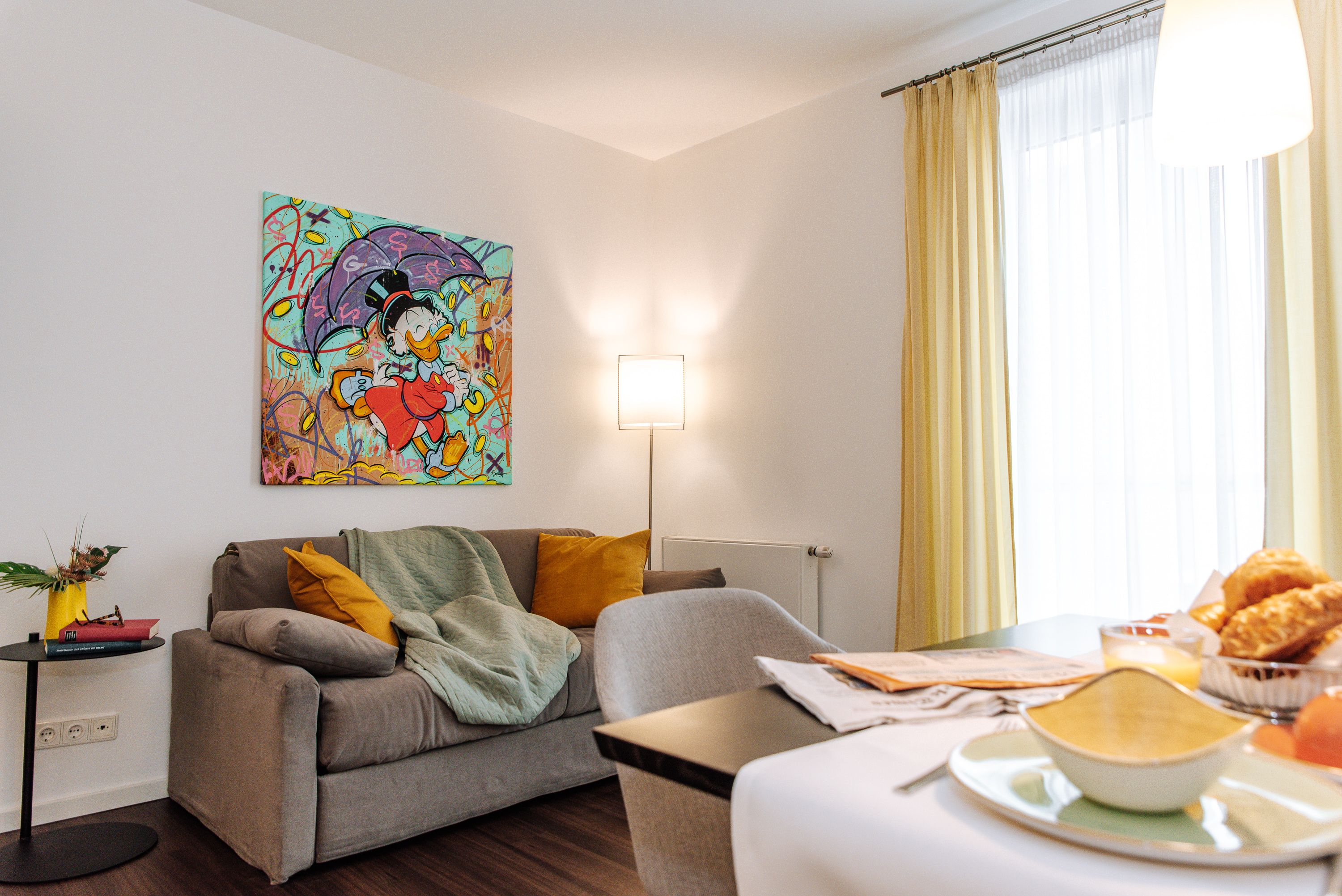 Living room with gray sofa, colorful comic art on the wall, yellow curtains and breakfast table.
