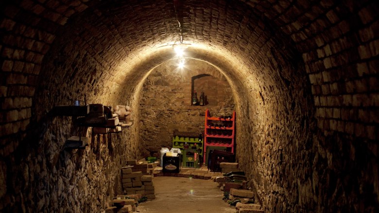 A narrow, vaulted wine cellar with brick walls and stored bottles.