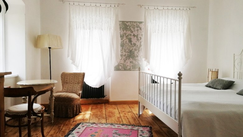 Room Mirandolina, © Gröger Bright room with wooden floor, white bed, two windows with curtains, a table and chair and a pink carpet.