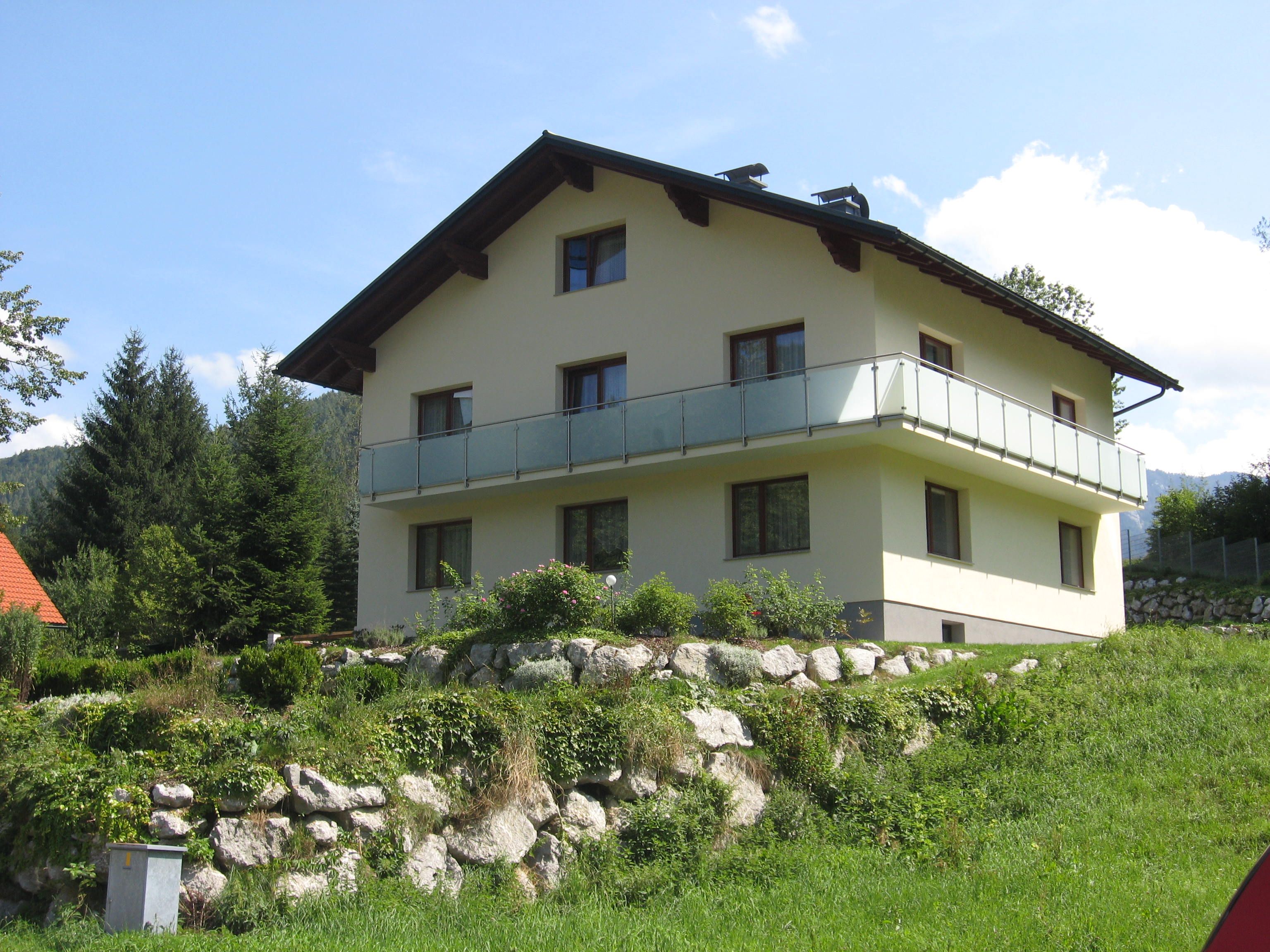 A two-storey house with balcony in a green, hilly landscape.