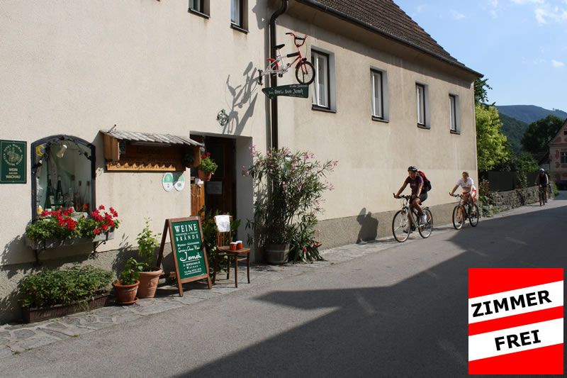 Exterior view of a house with bicycle decoration and wine bar. Two cyclists ride past. Sign with 'Room available'.