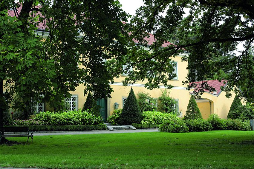 A yellow building with red roofs, surrounded by green trees and manicured lawns in a park.