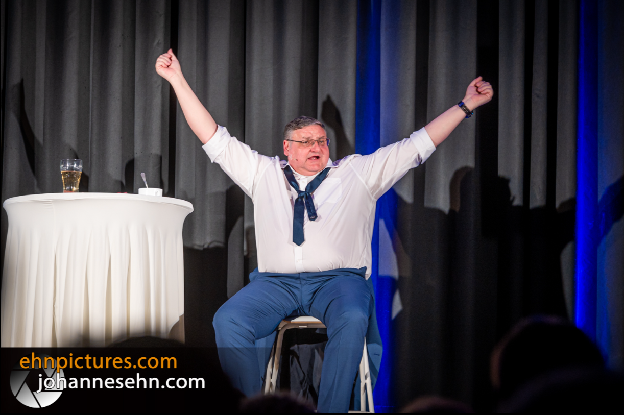 A man sits on a stage with his arms raised, next to him a table with a glass.
