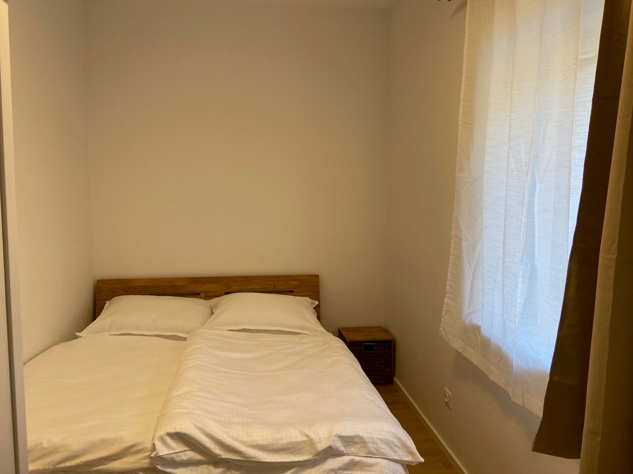 A simply furnished bedroom with a double bed, white sheets and a small bedside table. A window with a curtain is visible.
