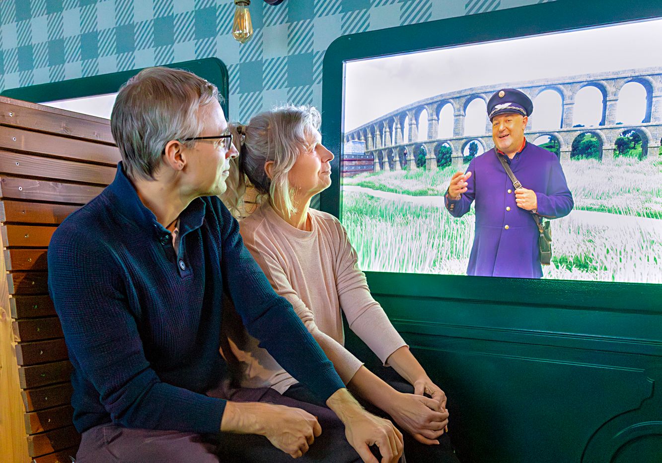 A couple is sitting in front of a screen showing a man in uniform in front of a viaduct.