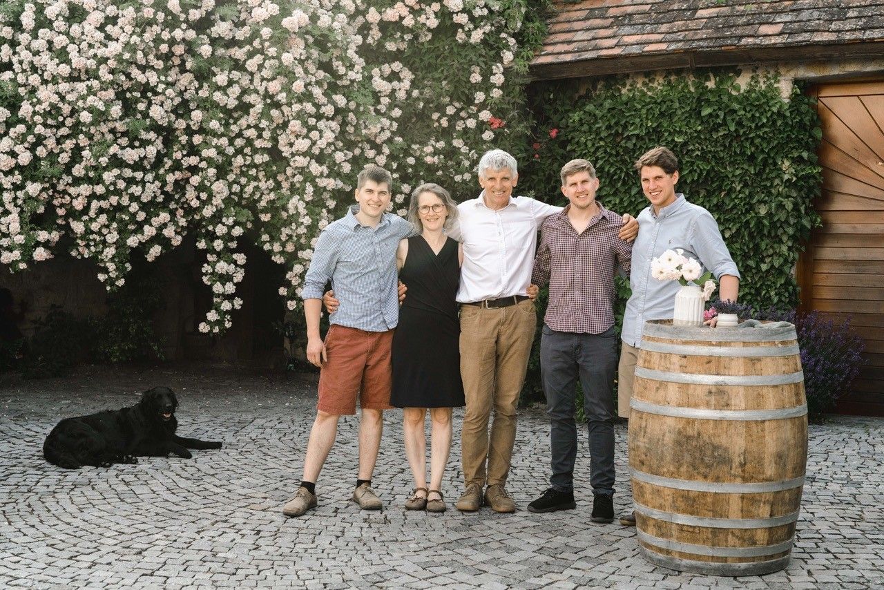 A family stands smiling in front of a flowering wall, with a dog in the background and a wine barrel with flowers on the right.