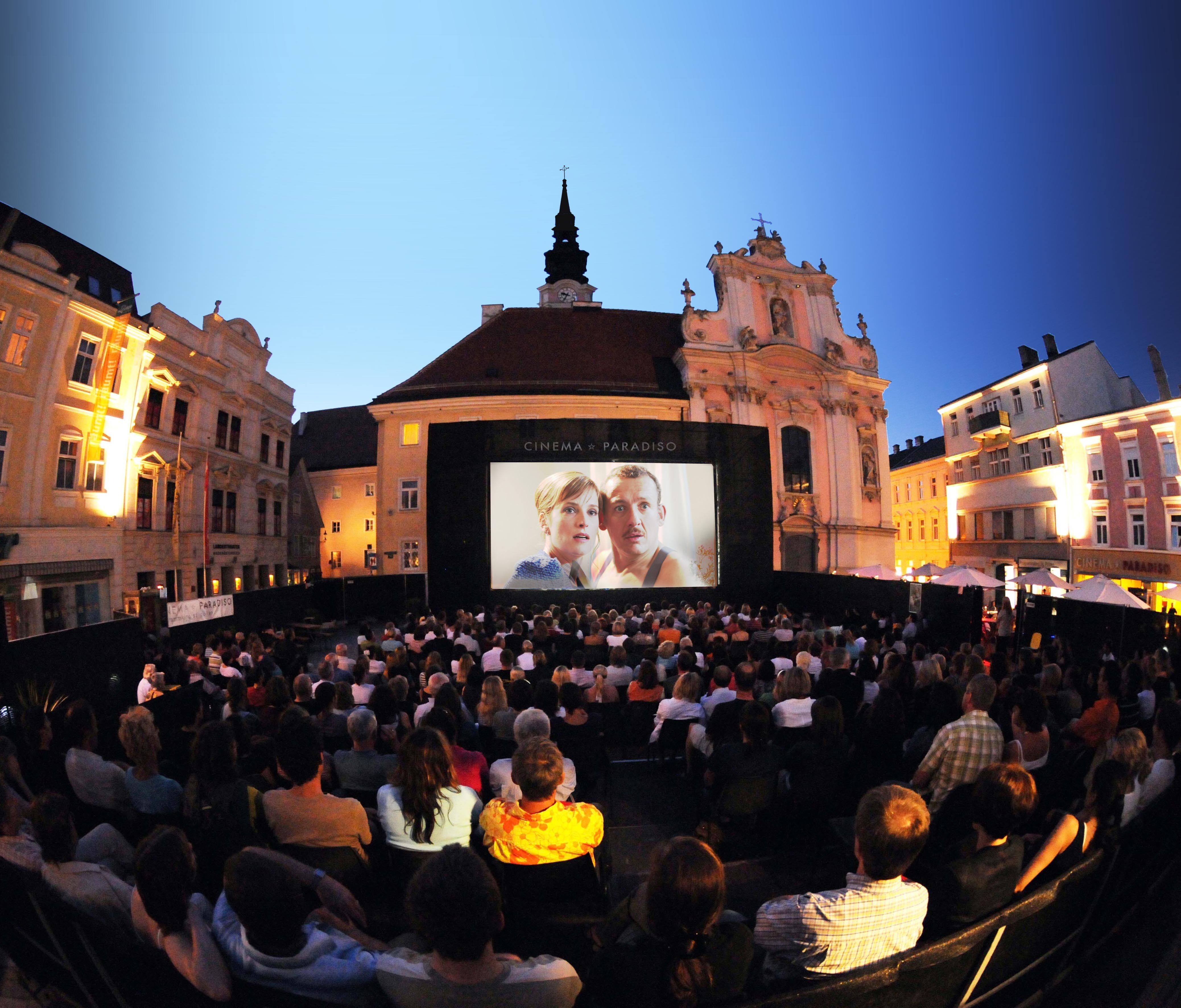 Open-air movie night in a square with a large audience in front of a big screen.