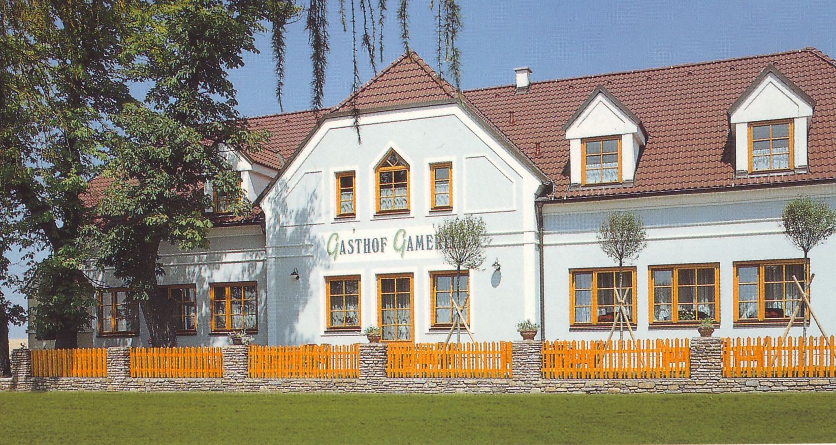 A traditional inn with a white façade and red roof tiles, surrounded by trees and a wooden fence.