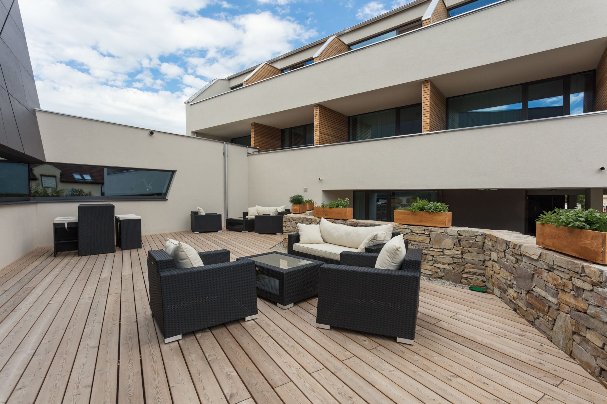 Modern terrace with wooden floor, black rattan furniture and stone wall, surrounded by a modern building.