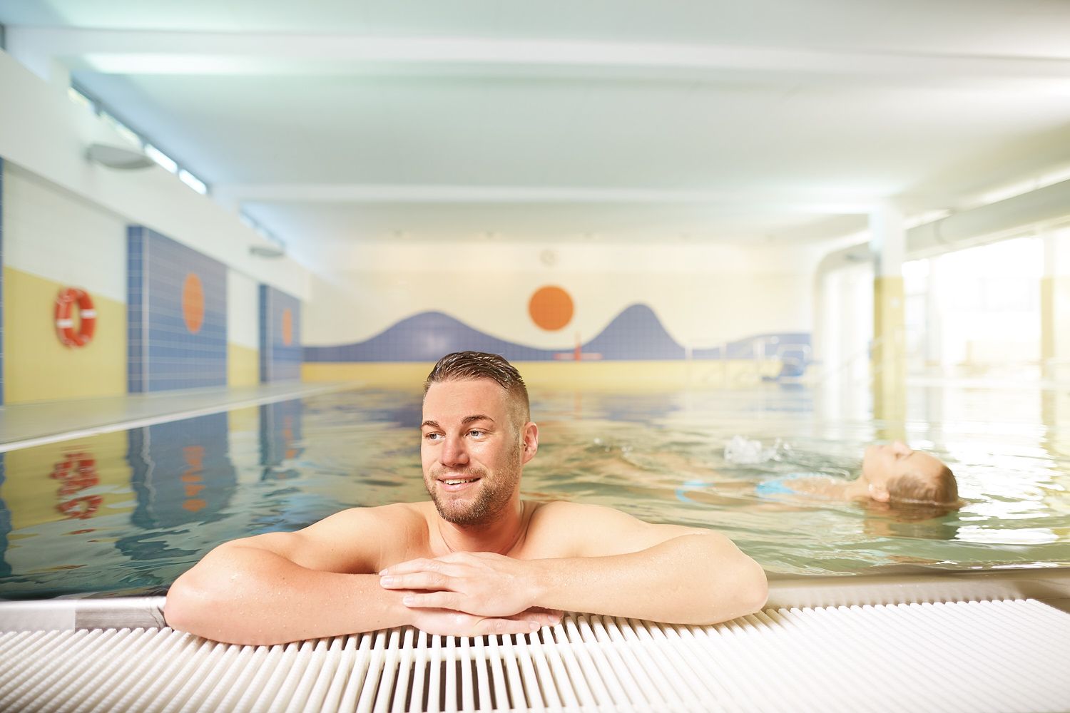 A man leans against the edge of an indoor swimming pool while a person swims in the water in the background.
