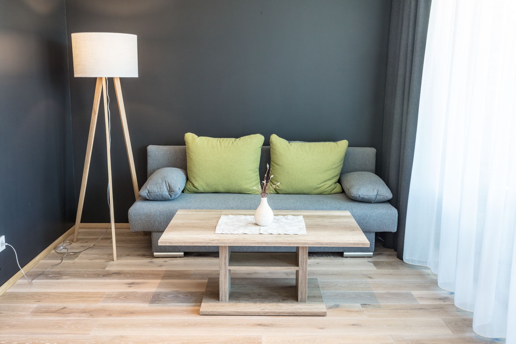 Modern living room with gray sofa, green cushions, wooden lamp and coffee table in front of a dark wall.