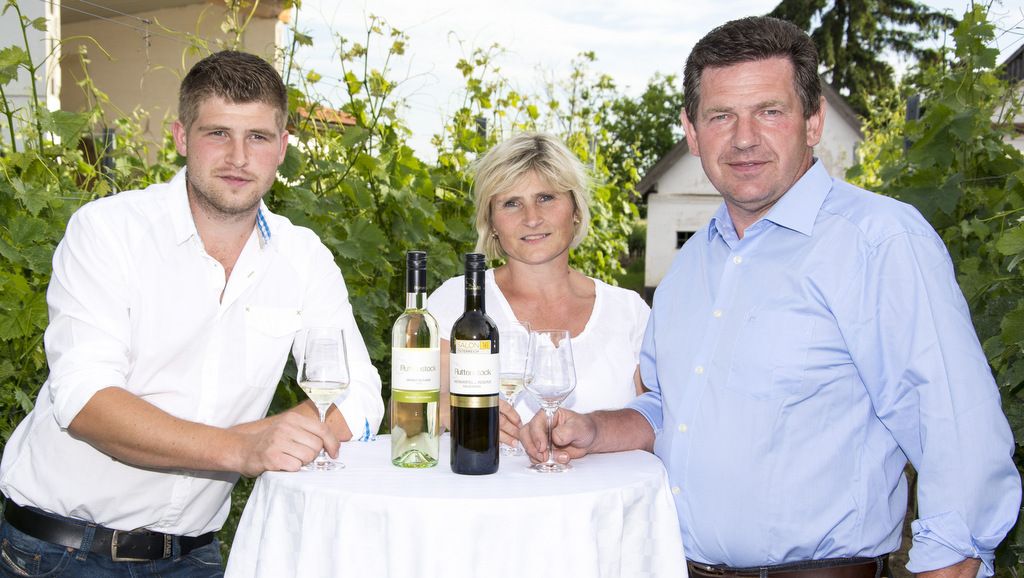Three people are standing at a table with bottles of wine outside.