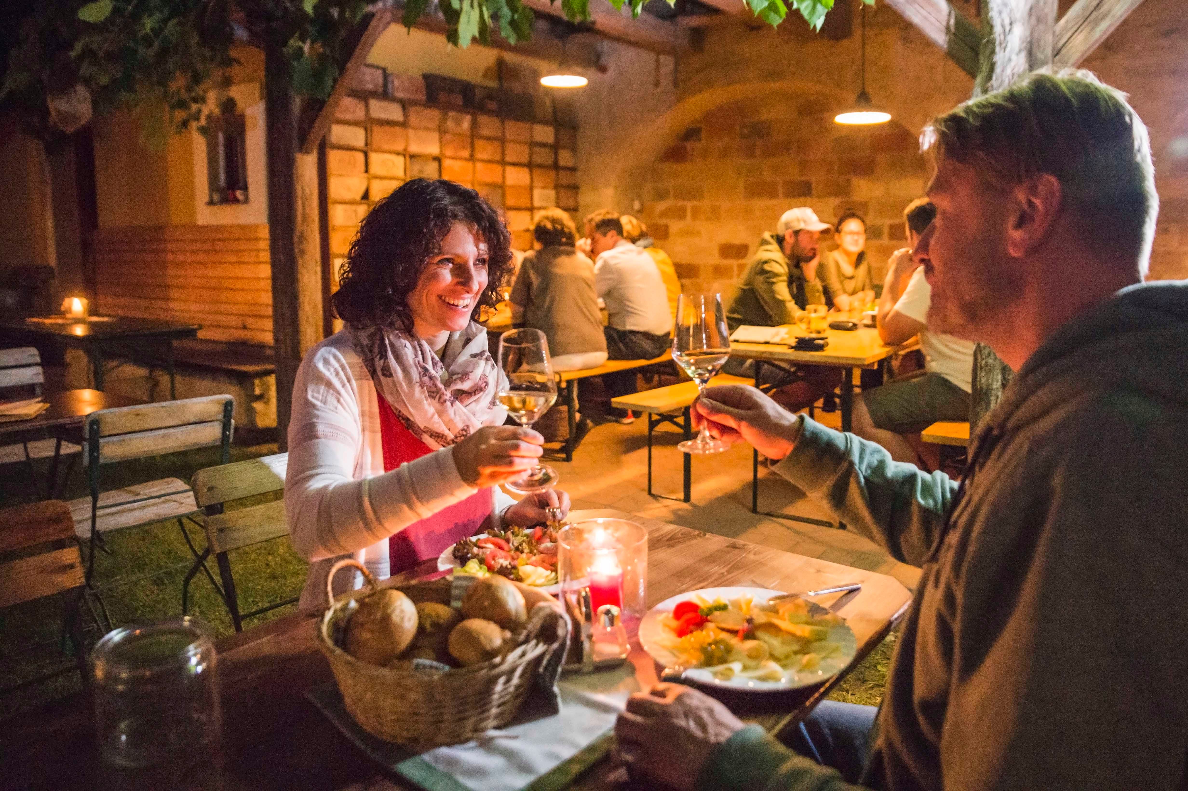 A couple toast with glasses of wine at a wine tavern, with other guests sitting at wooden tables in the background.