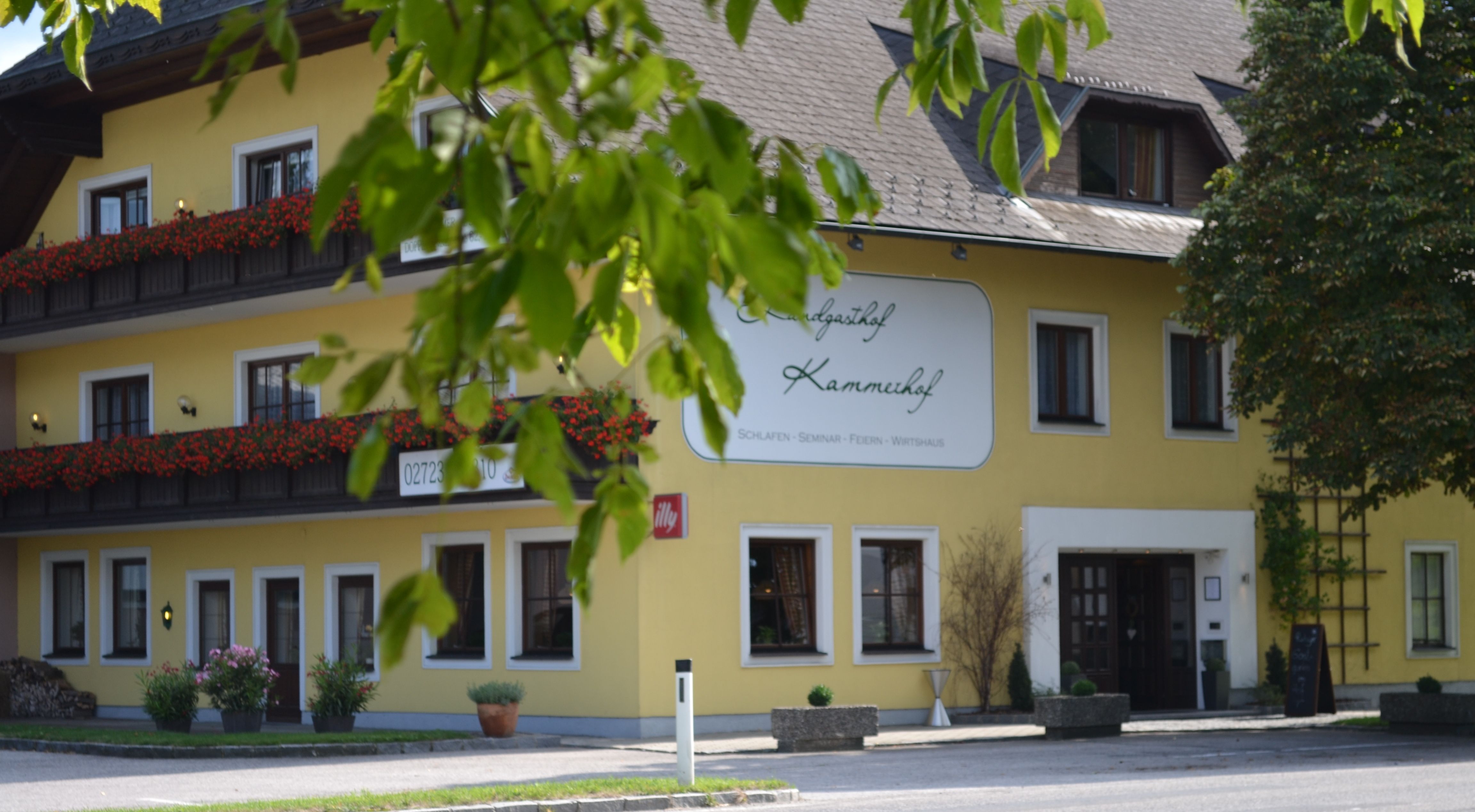 Yellow building with sign 'Landgasthof Kammerhof', flower boxes and trees in the foreground.