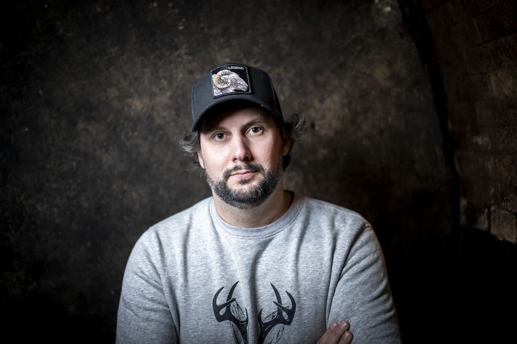 A man with a beard and baseball cap stands in front of a dark wall.