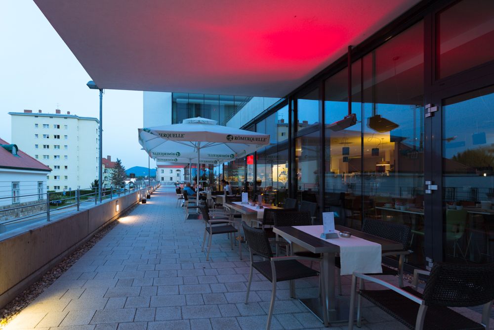 Terrace of a restaurant with tables and chairs, illuminated by red lights, with a view of buildings in the background.