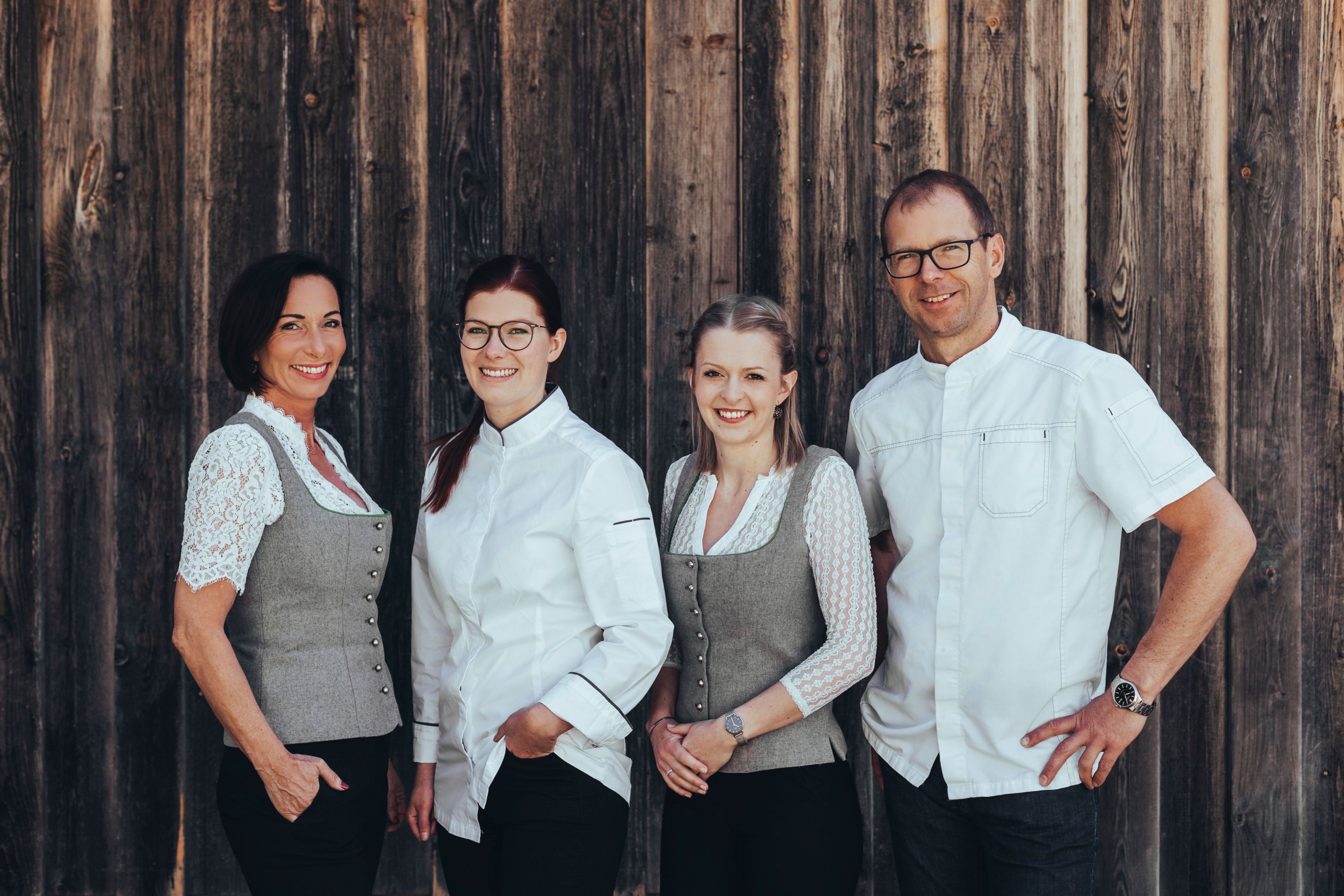 Four people are standing in front of a wooden wall, two women in traditional costume and two people in white clothing.