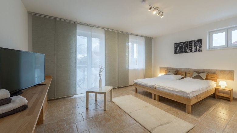 Modern bedroom with double bed, TV, table and large windows with curtains.