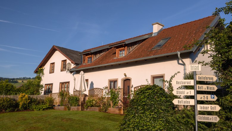 A white building with a red roof and several signs in the foreground, surrounded by a green landscape.