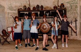Group of children in medieval helmets and with weapons posing in a room with armor.