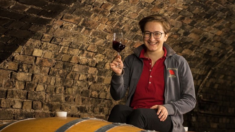 A person sits on a wine barrel in a wine cellar and holds a glass of red wine in his hand.