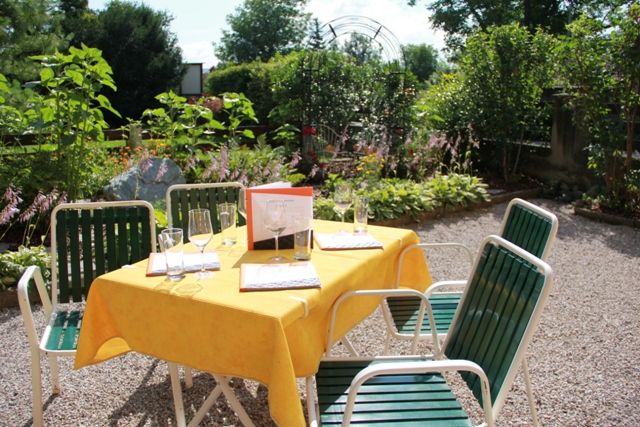 A sunny guest garden with a table set with a yellow tablecloth and glasses, surrounded by green plants.