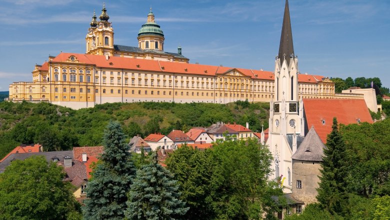 View of Melk Abbey and the parish church in Melk, surrounded by green countryside and blue sky.