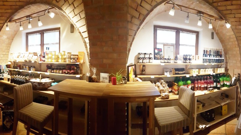Interior view of a small store with shelves full of food and drinks, wooden table and chairs.