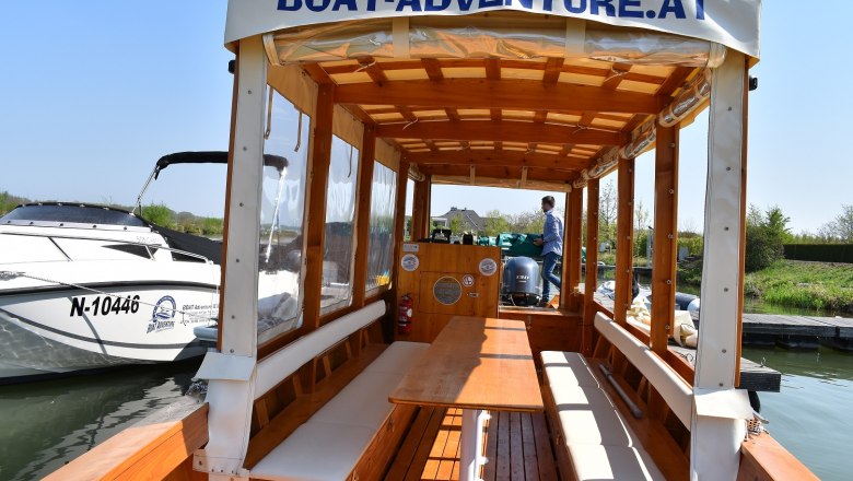Interior view of a wooden boat with benches and table, in the background a man at the helm.