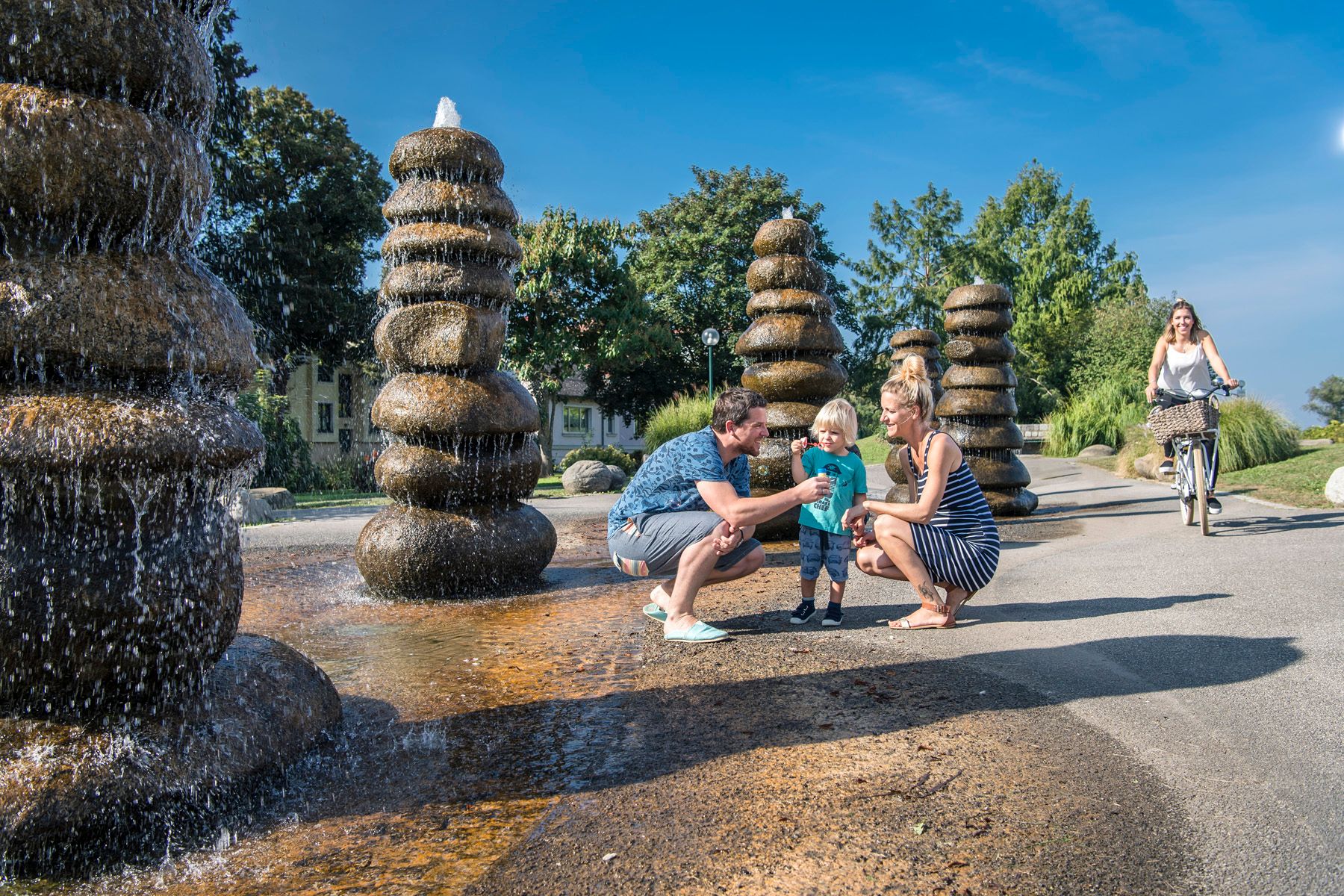 Family at a fountain in a park, with a woman on a bicycle in the background.