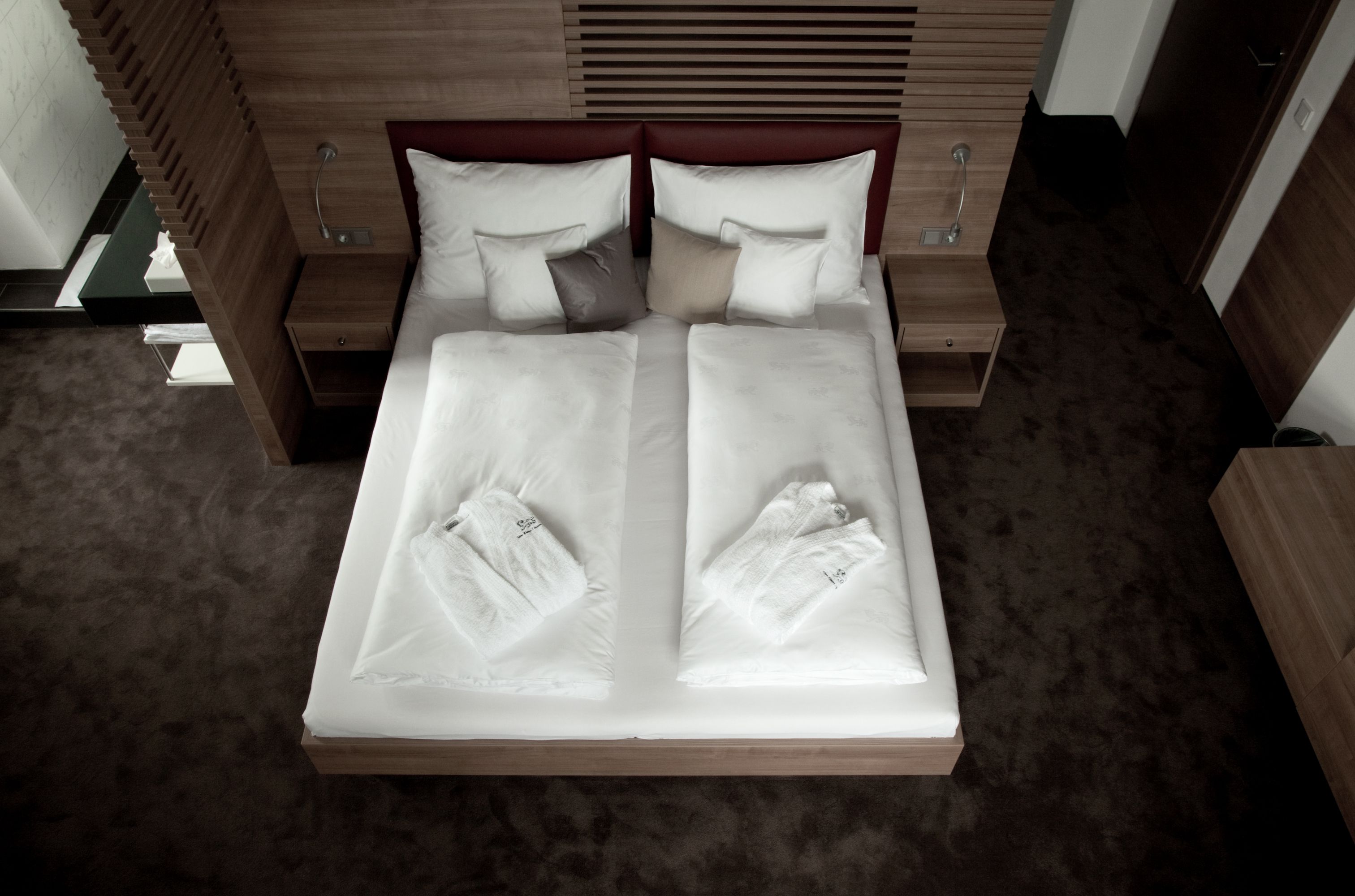 A modern hotel room with a double bed, white comforters and bathrobes, surrounded by wooden furniture.
