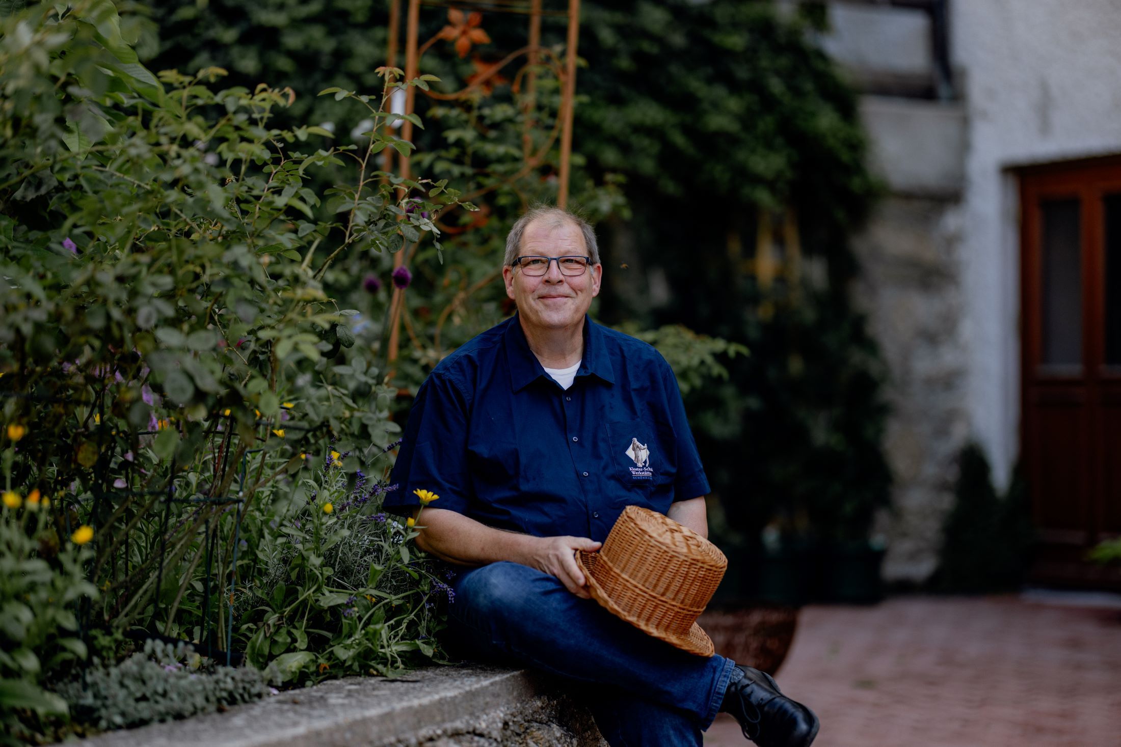 A man sits in a garden and holds a woven hat.