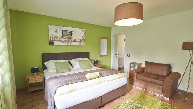 Modern furnished guest room with green accent, double bed, brown armchair and decorative picture on the wall.