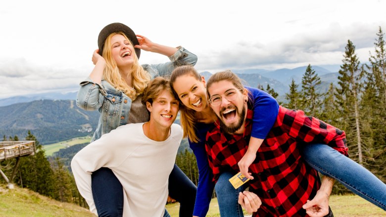 Four smiling people in a mountain landscape, two carrying the others on their backs.
