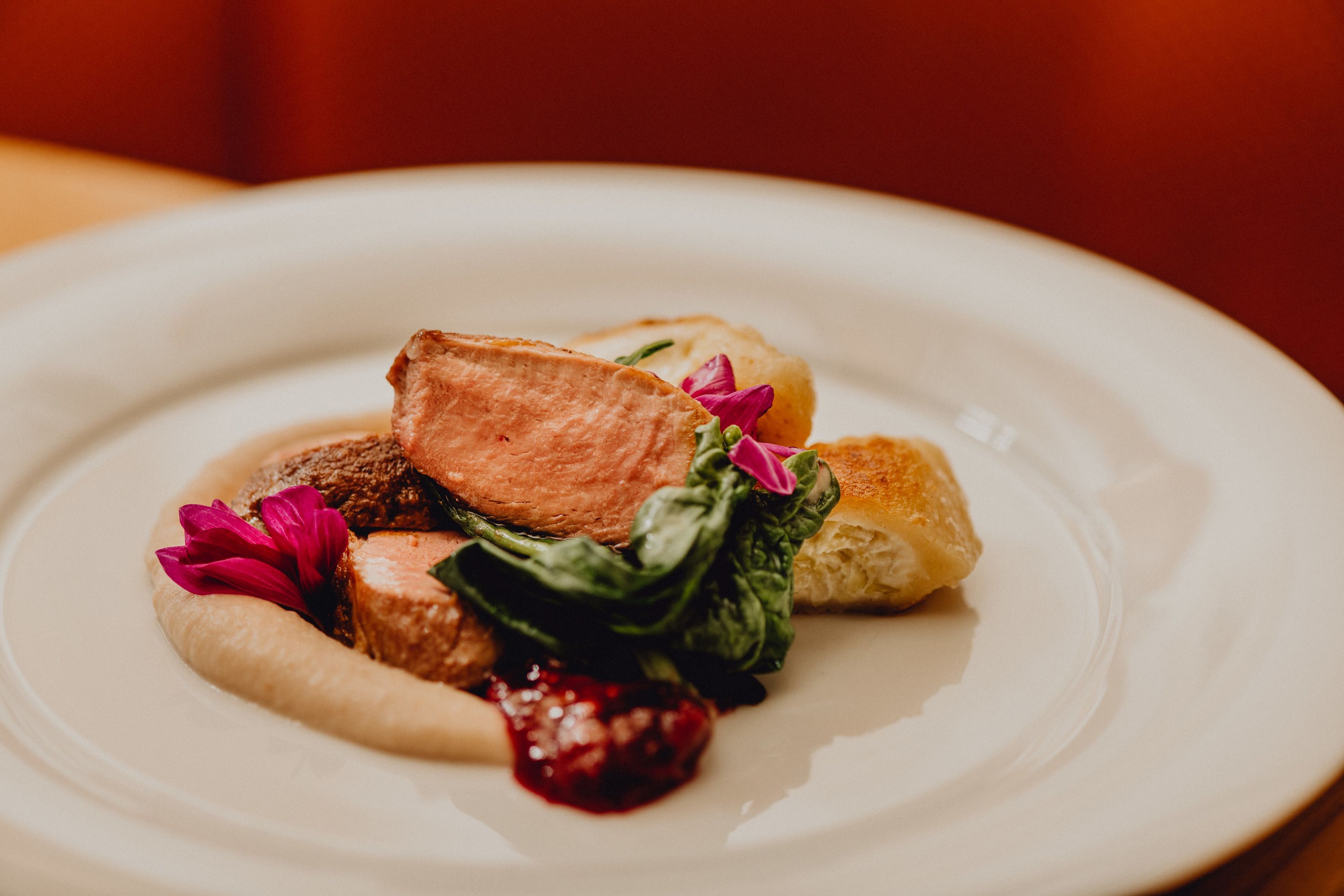 A plate of pink roast duck, cream tartlets, chestnut purée and green leaves, garnished with edible flowers.
