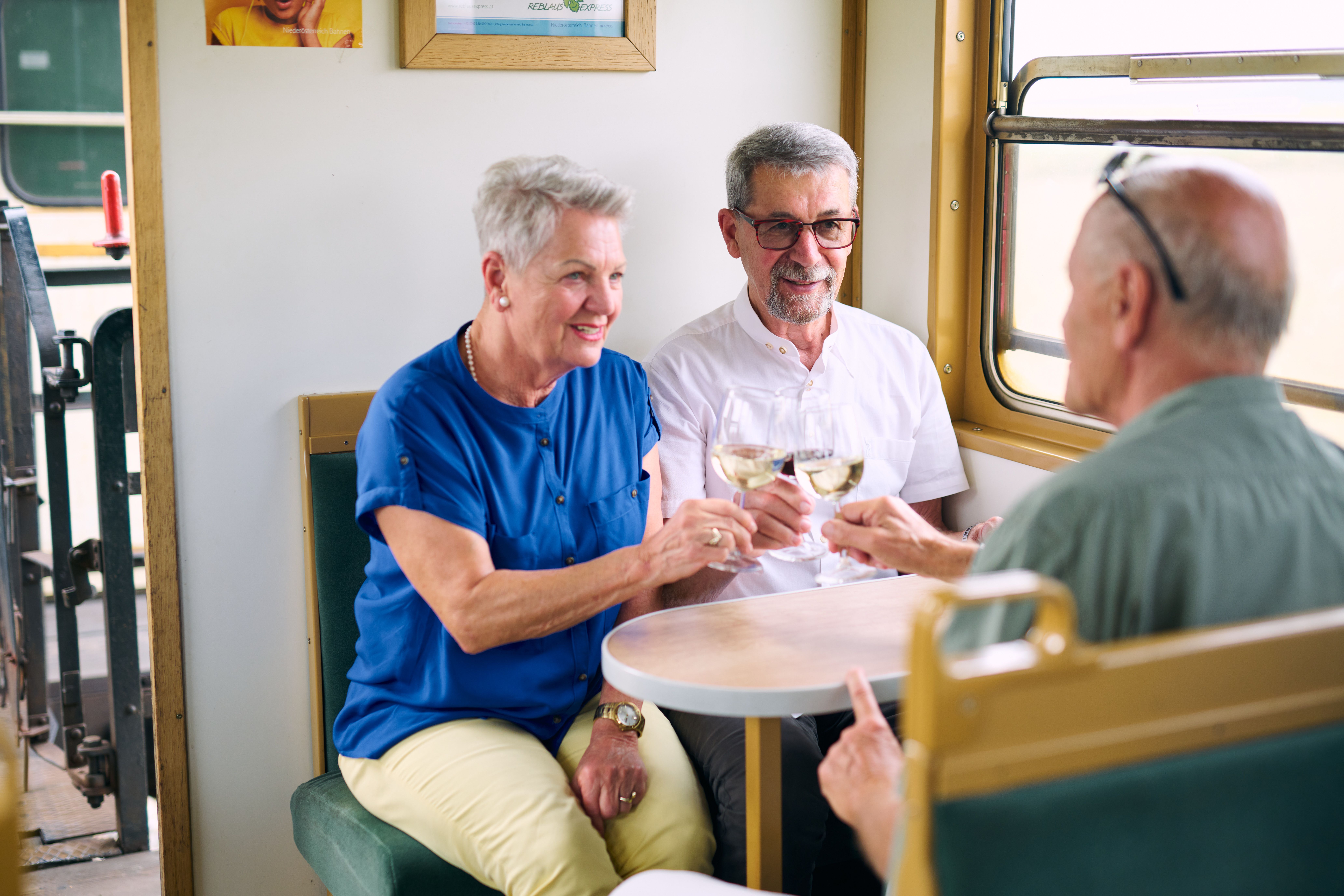 Three elderly people clink glasses of wine in a train compartment.
