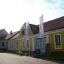 Yellow building with red roof tiles and two flags in front of it.