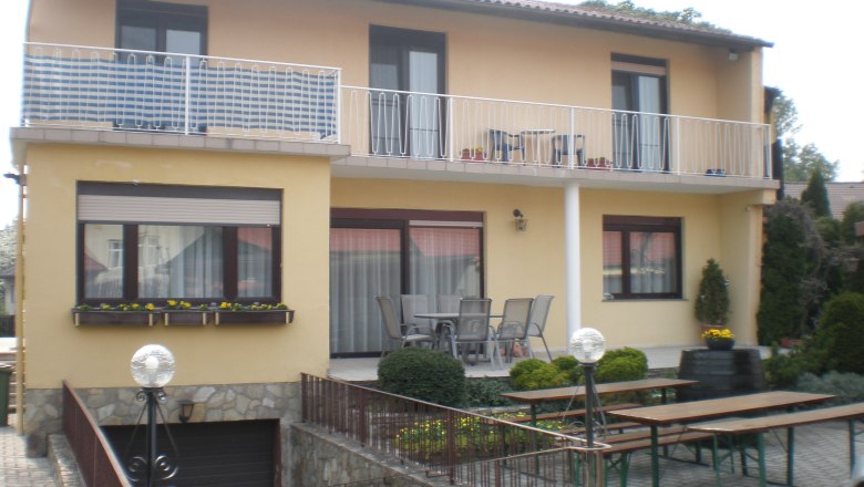 Two-storey house with balcony and terrace, surrounded by garden furniture and plants.