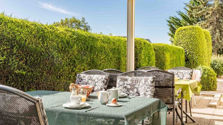 Outdoor breakfast table with crockery and green tablecloth, surrounded by hedges.