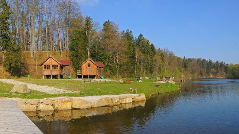 Two wooden huts on the shore of a lake, surrounded by trees and green grass.