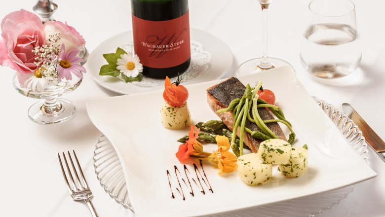 Elegant dish with fish, asparagus and potatoes on a white plate, decorated with flowers. A bottle of wine and a glass in the background.