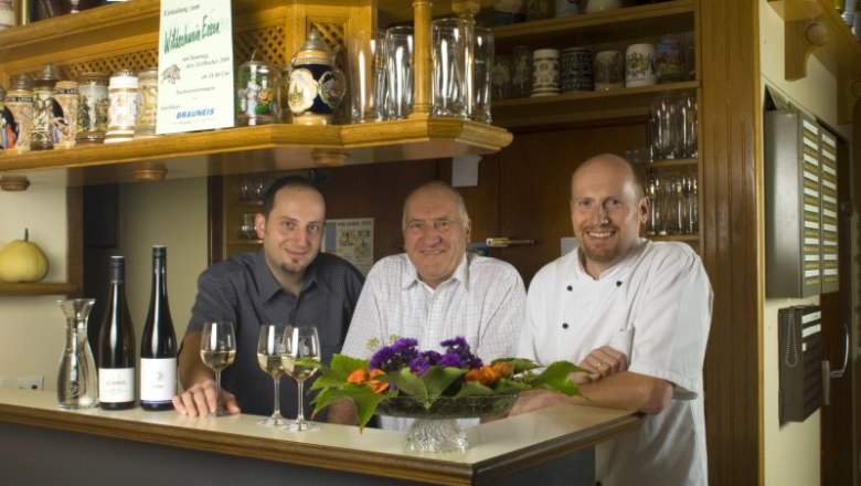 Three men are standing behind a bar with bottles of wine and glasses.
