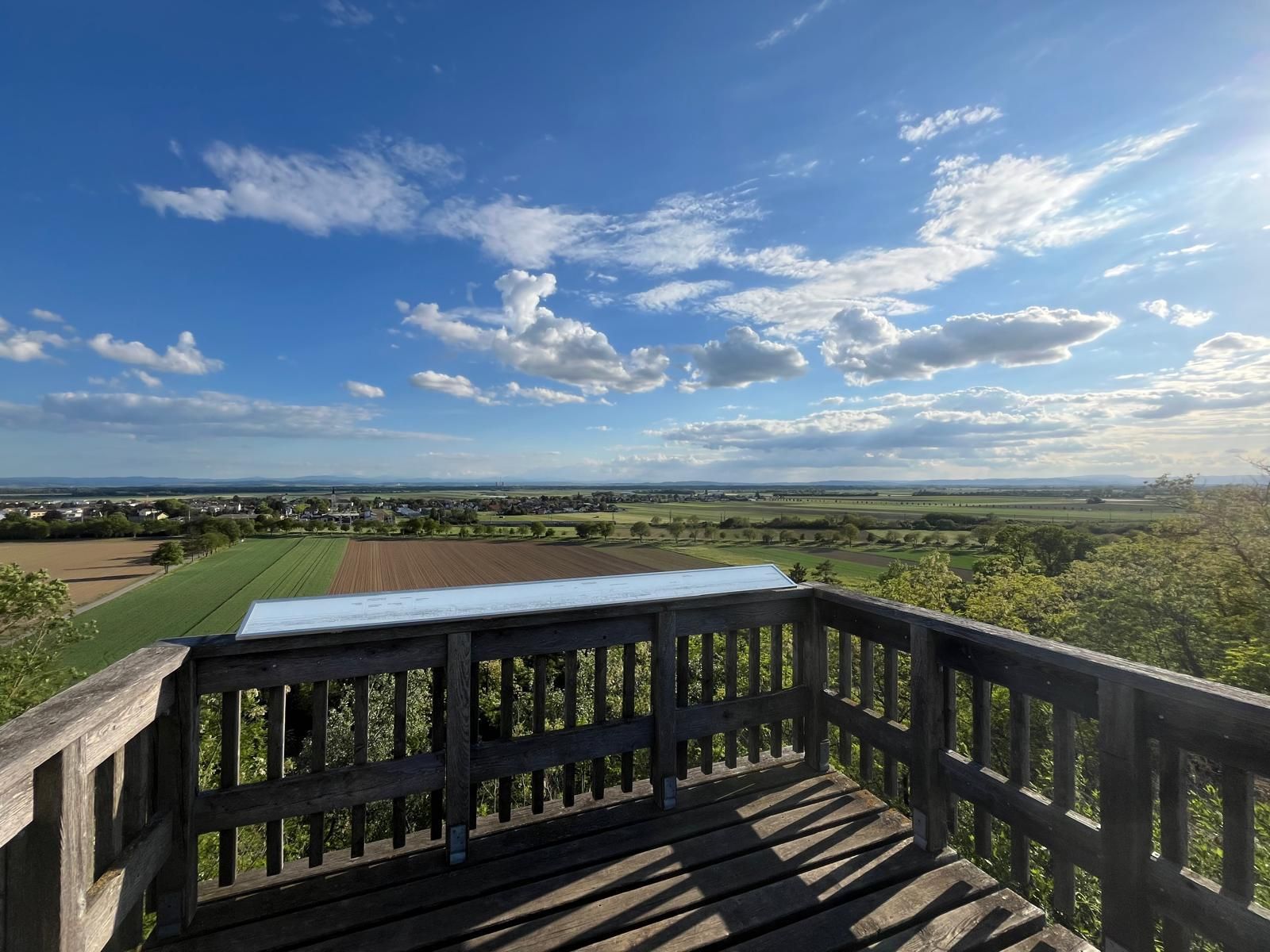Wooden viewing platform with railings above a vast landscape of fields and small villages. Above it stretches a large blue sky with numerous white clouds, illuminated by the low sun. The scene conveys vastness and tranquillity.