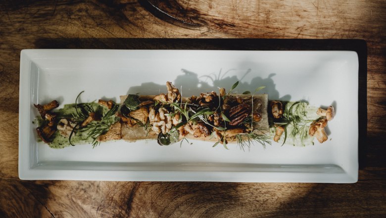 A rectangular plate with sautéed chanterelles, pecan nuts, wild herbs and root bread, artfully arranged on a wooden table.