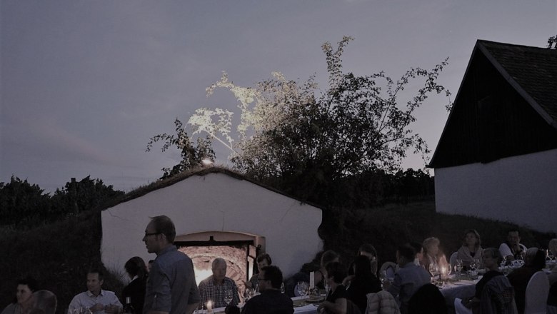 People sit at a long table outside at dusk, surrounded by buildings and trees.